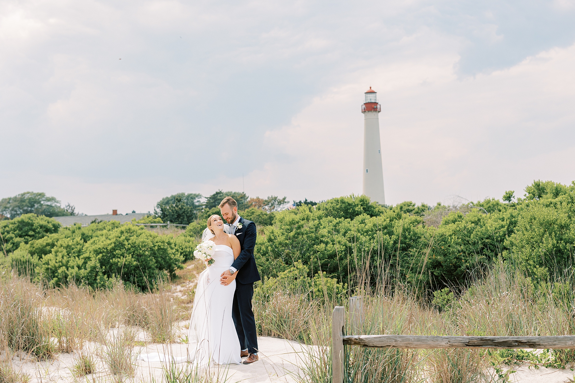bride and groom hug in front of Cape May lighthouse on beach