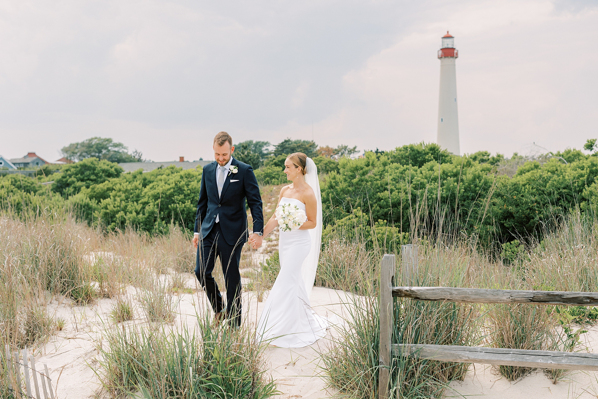 bride and groom hold hands walking on beach in front of Cape May lighthouse 