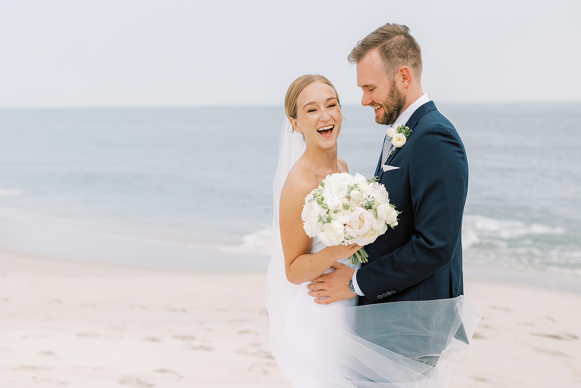 groom looks down at bride making her laugh with veil around them on Cape May beach