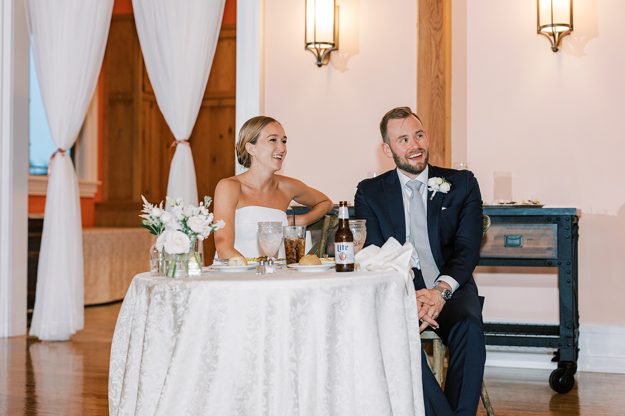 bride and groom laugh sitting at table listening to toasts 