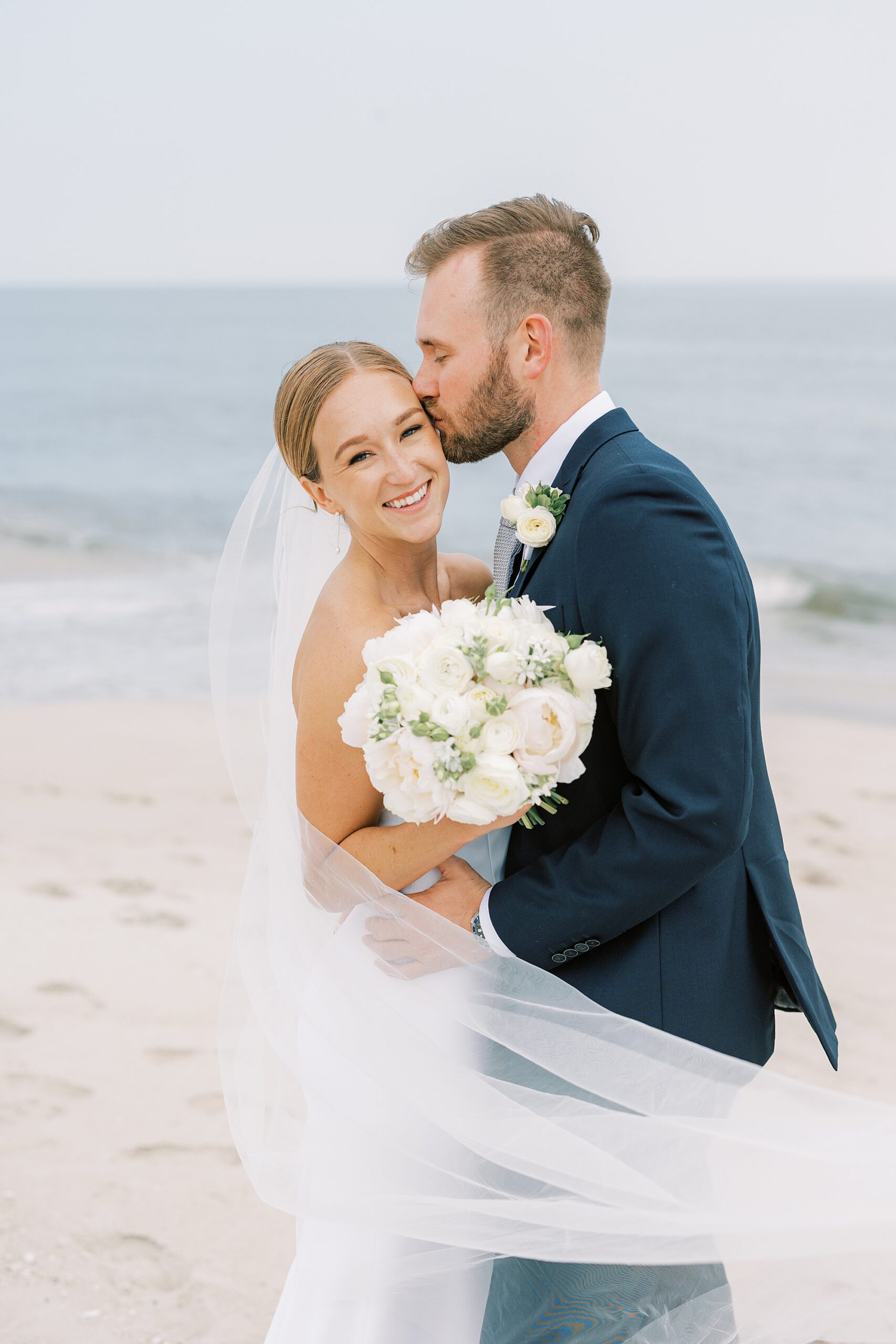 groom leans to kiss bride's forehead holding ivory bouquet on beach in Cape May NJ