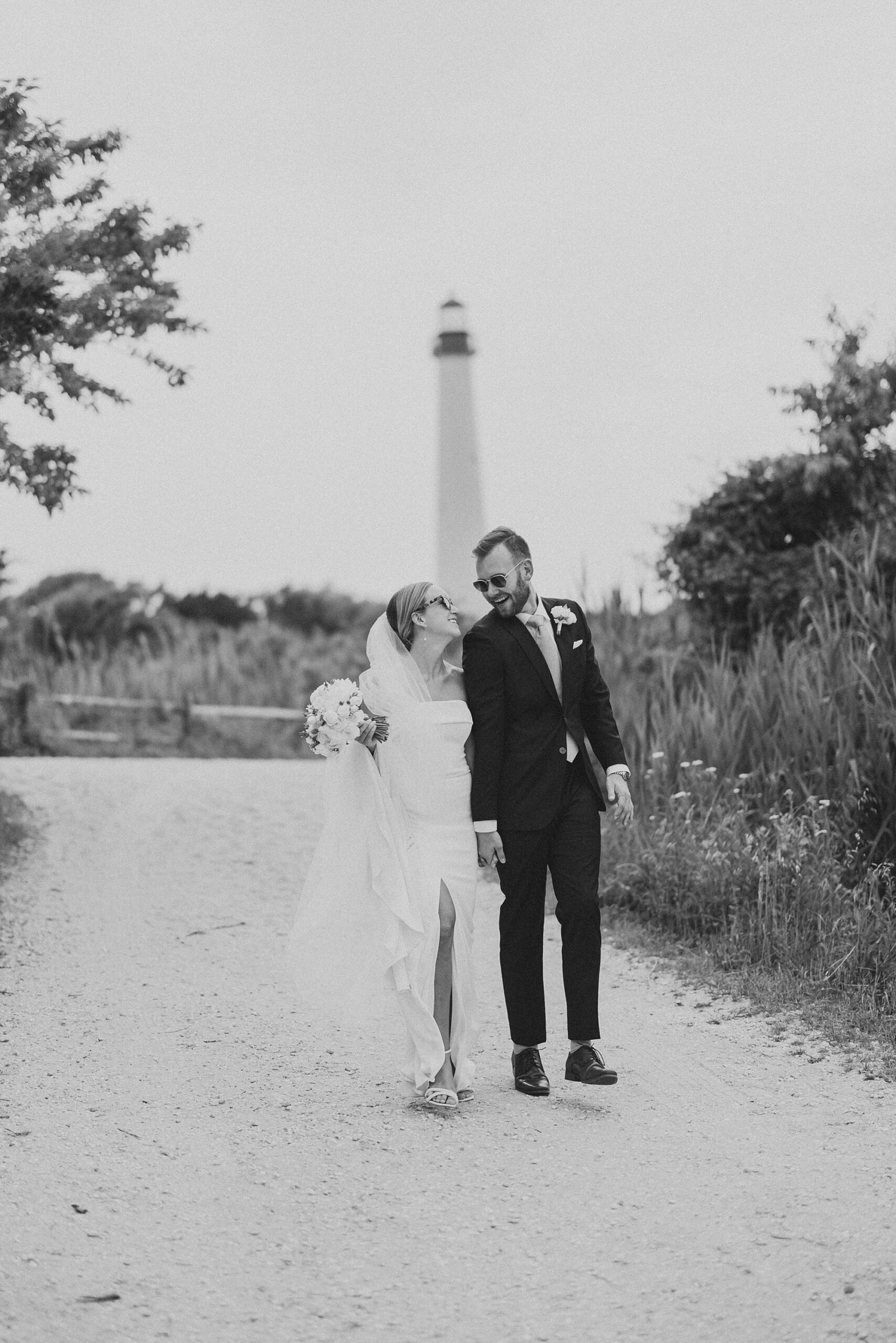 bride and groom hold hands walking in front of Cape may lighthouse wearing sunglasses 