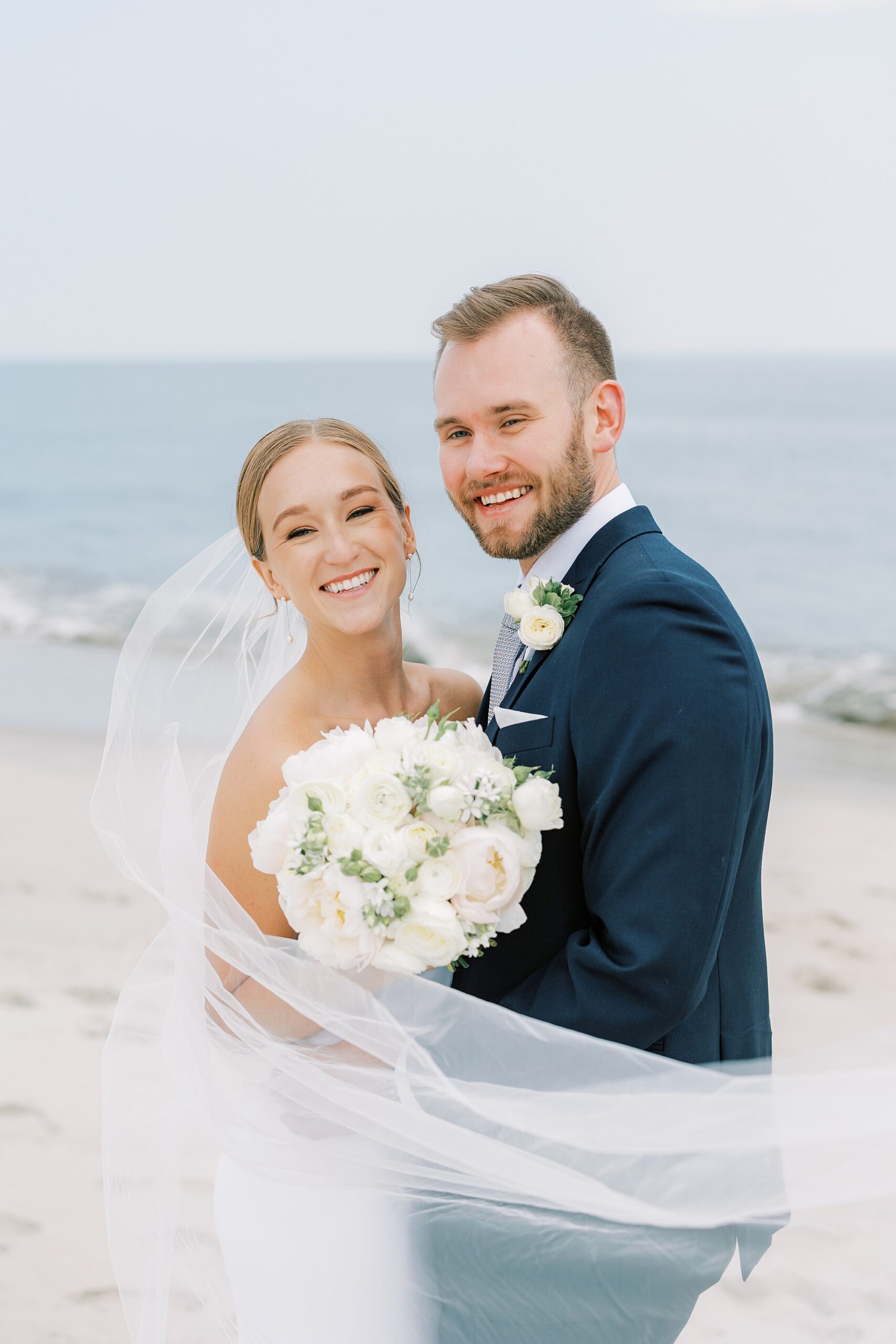bride and groom hug on beach with veil floating around them