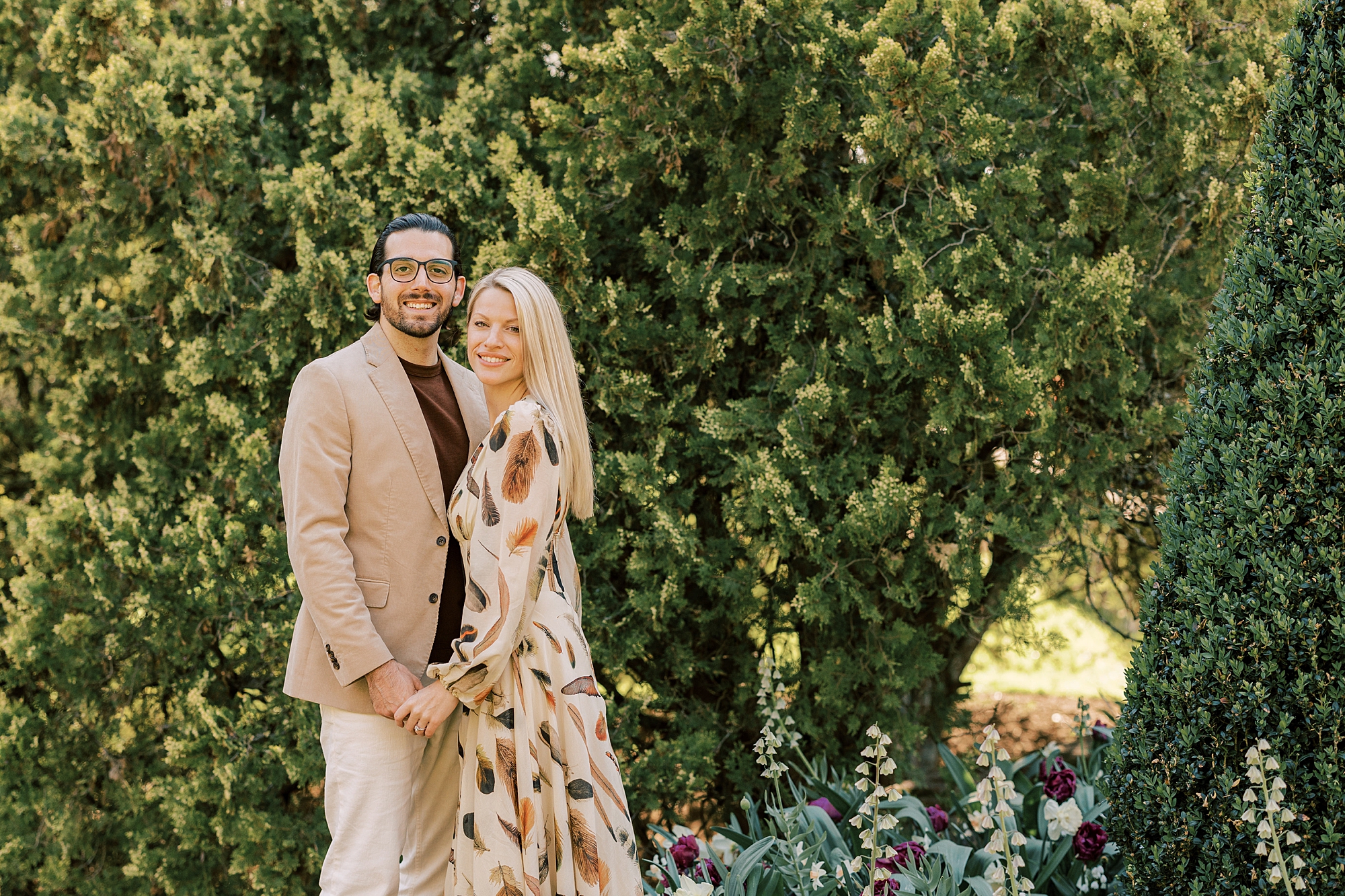man and woman hold hands in front of green trees at Longwood Gardens 