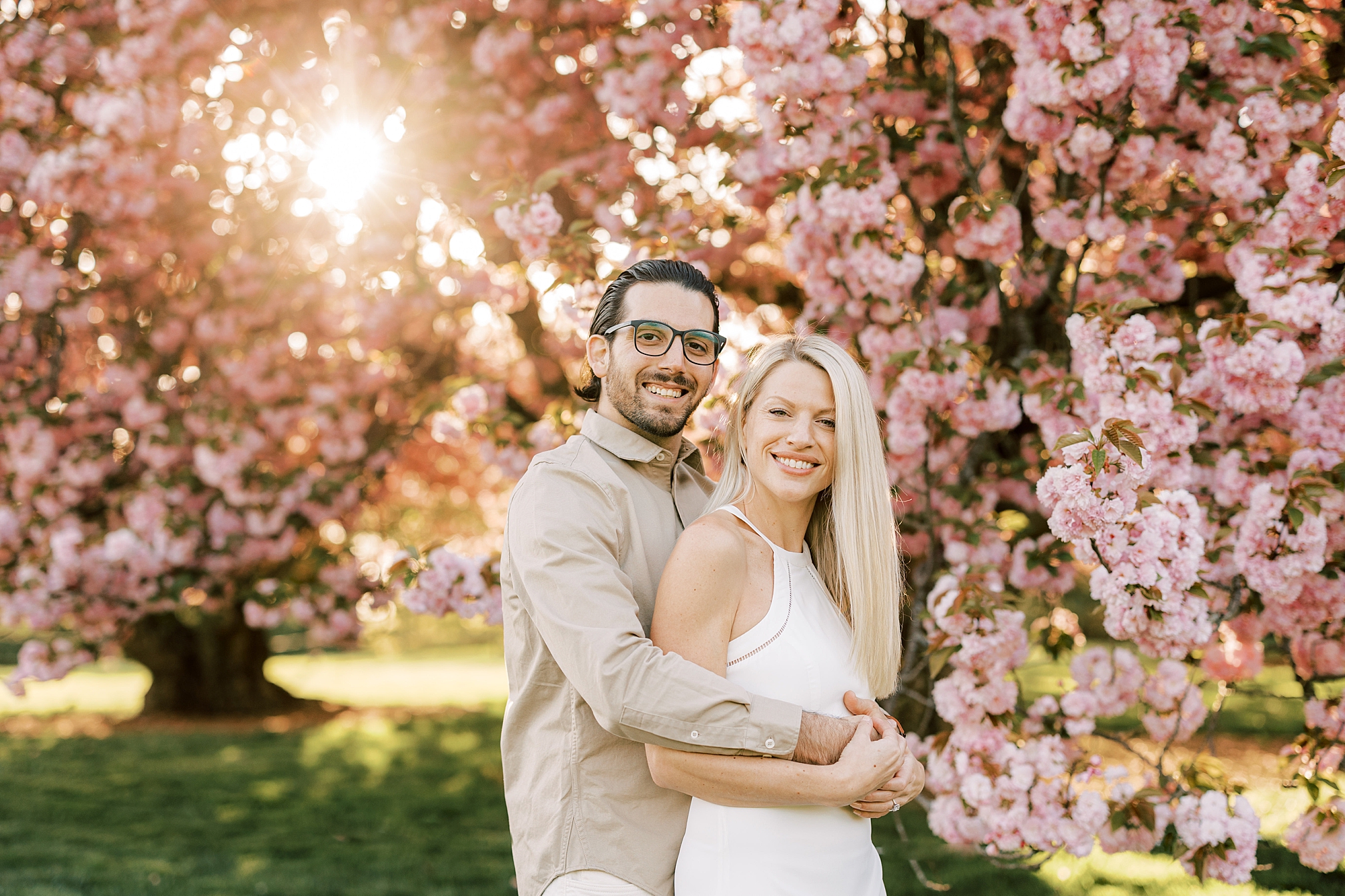 man hugs woman from behind in front of cherry blossom trees