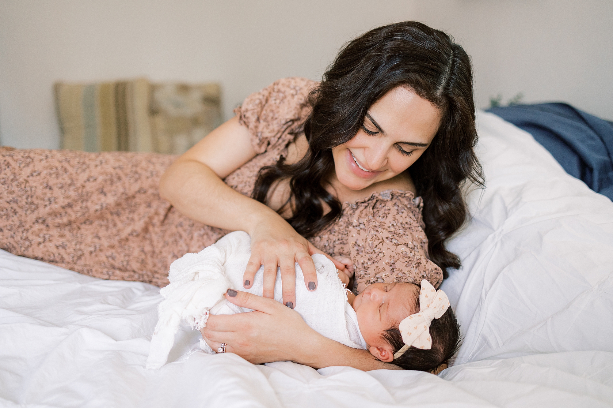 mom leans on bed with newborn daughter in pink bow