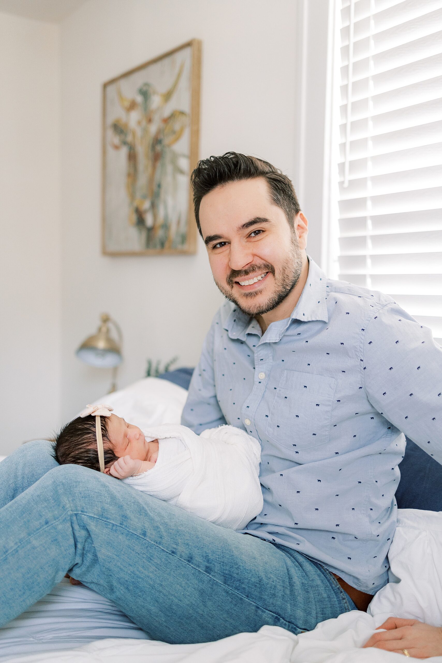man in blue shirt holds newborn daughter on lap during newborn portraits at home in Media PA