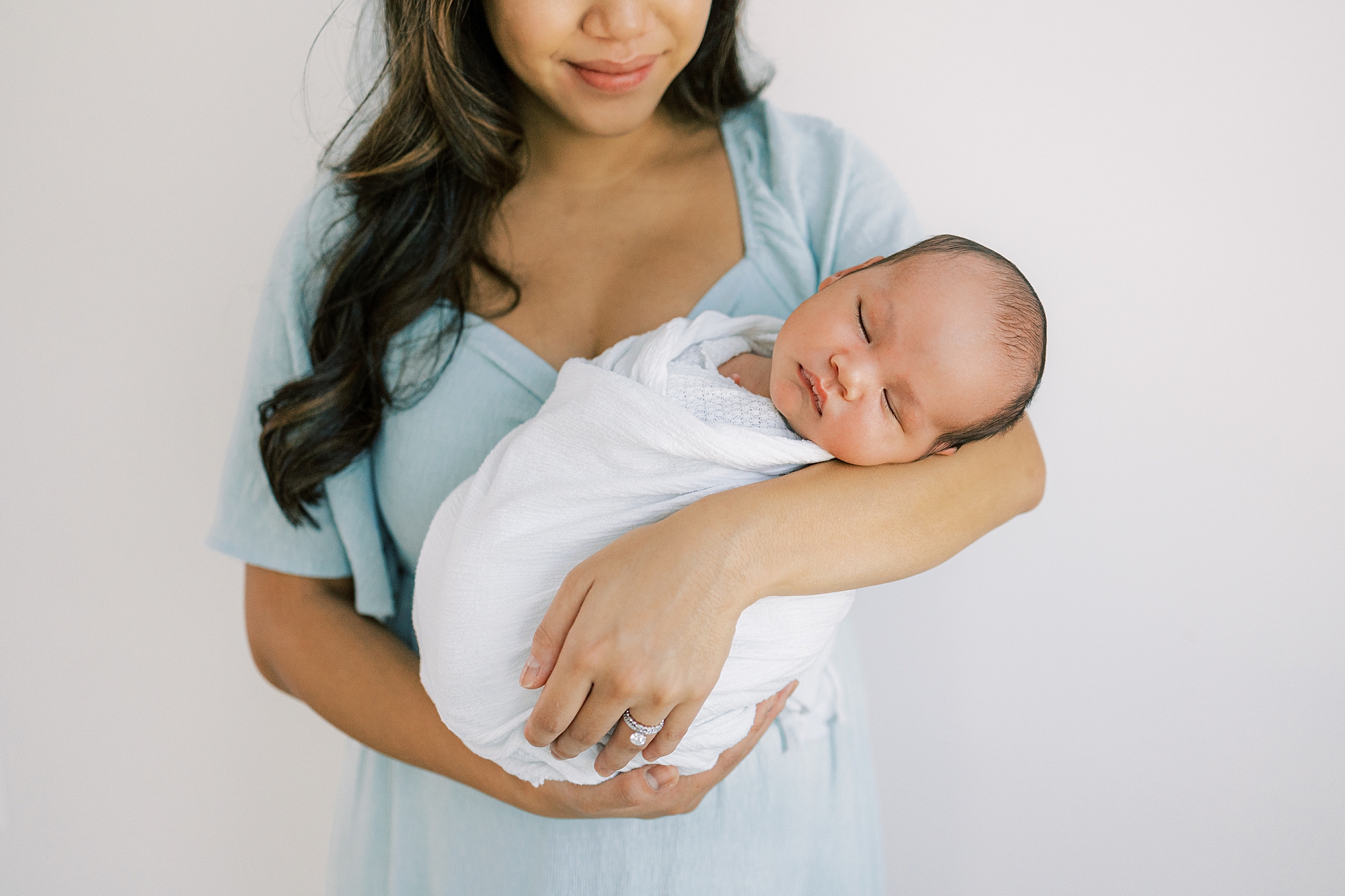 mother in blue dress holds baby boy in white wrap during newborn session in Cherry Hill NJ