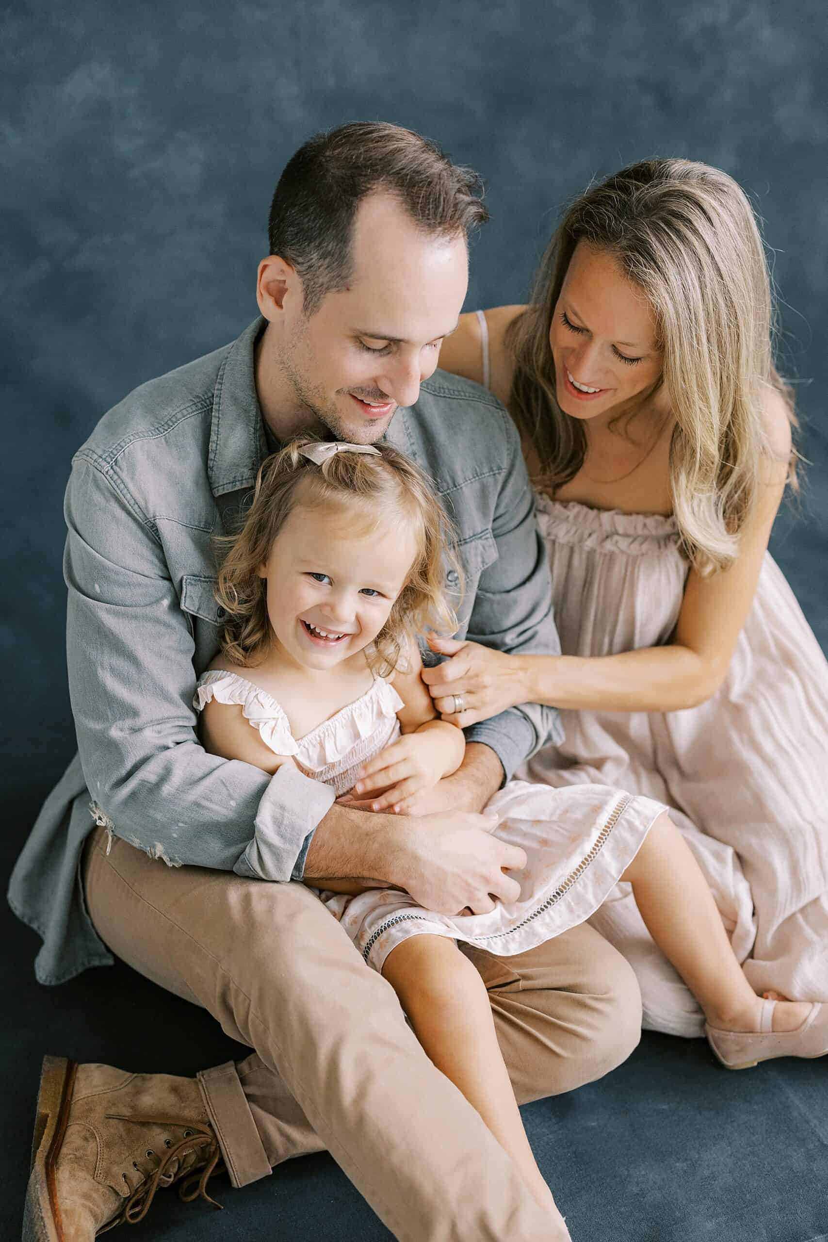 a stylish father sits on a black painted backdrop holding his 5 year old daughter in his arms while she giggles and his wife holds on to his left arm and smiles at their daughter