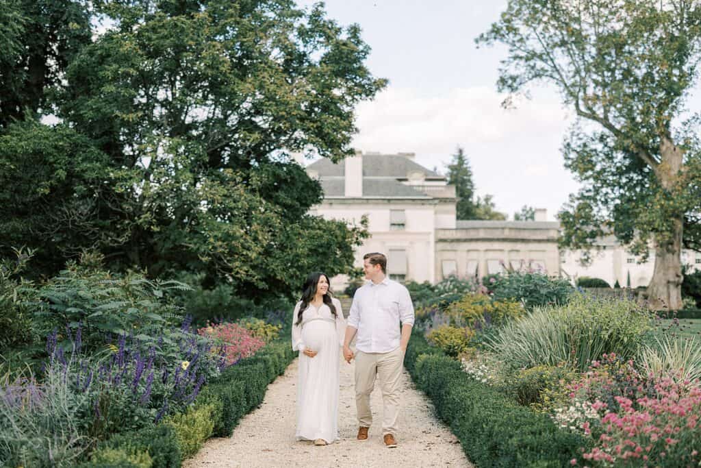 a pregnant woman smile at her husband as they hold hands and walk through the floral gardens at Nemours Mansion and Gardens in Wilmington DE a great alternative to Longwood Gardens for photo sessions