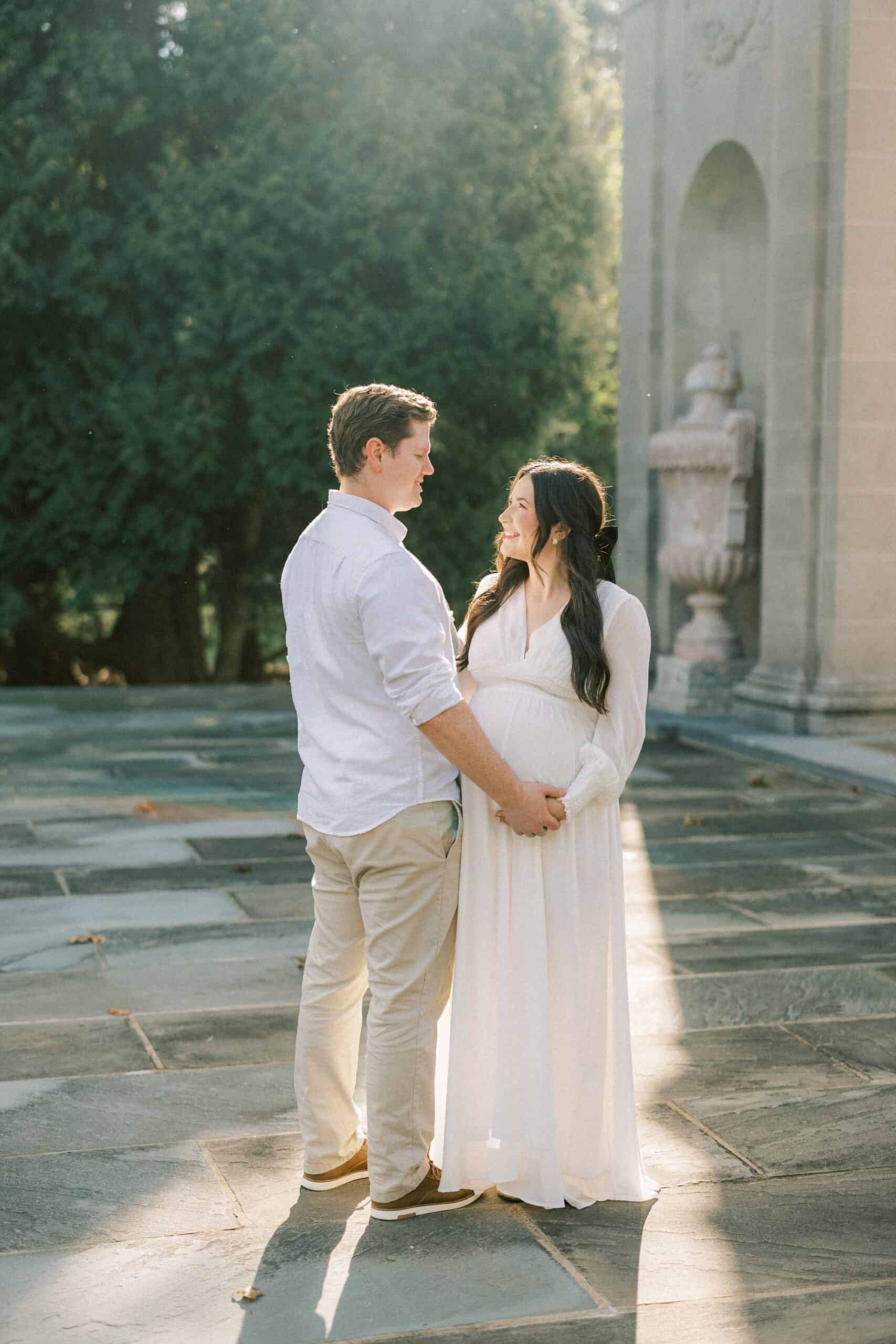 a married couple hold hands and smile at each other in the golden hour sunshine at Nemours Mansion and Gardens. the woman is pregnant in a long white maternity dress