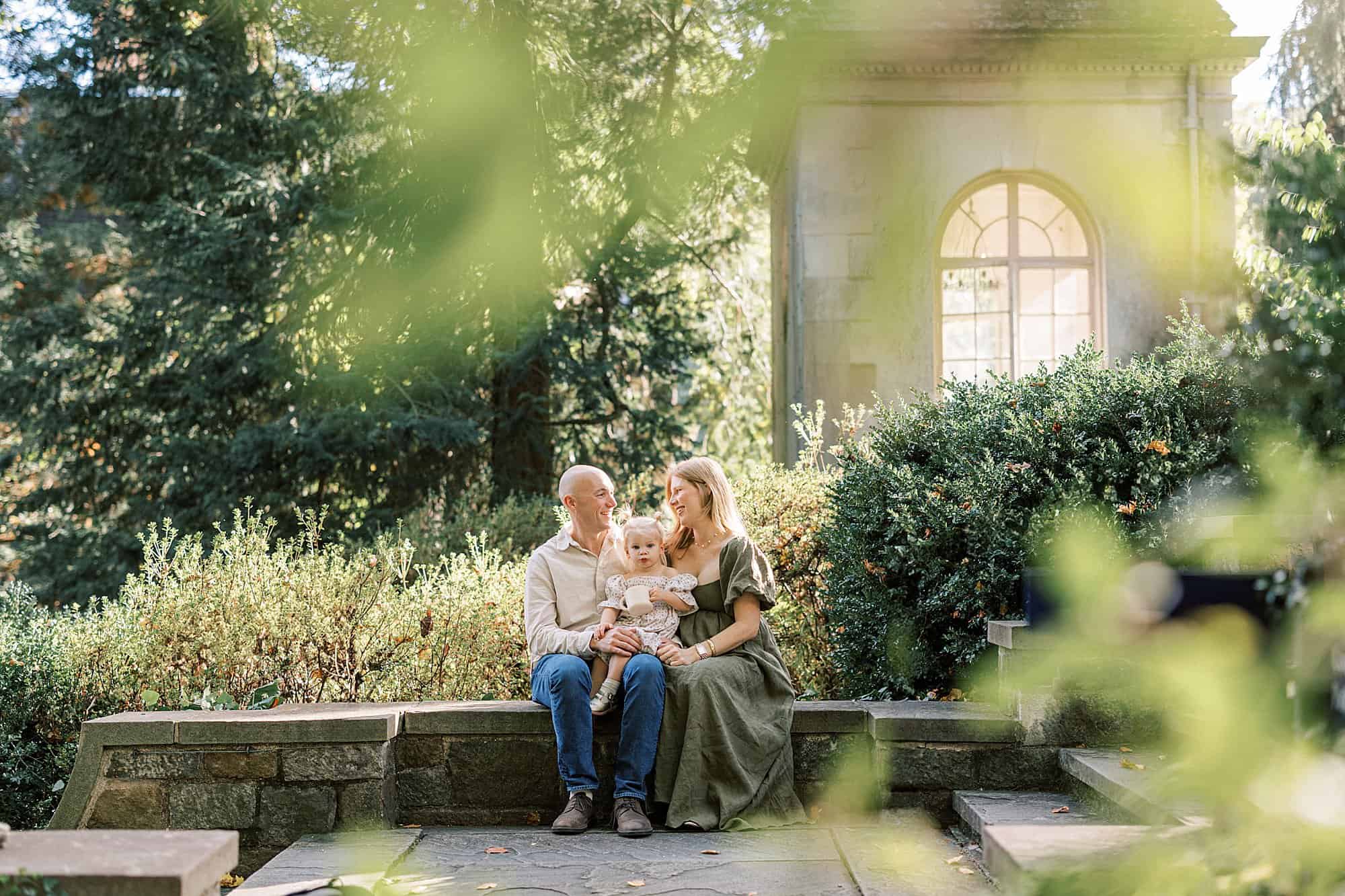 a pregnant mother and her husband sit with their 2 year old daugher in the Longwood Gardens adjacent Winterthur gardens