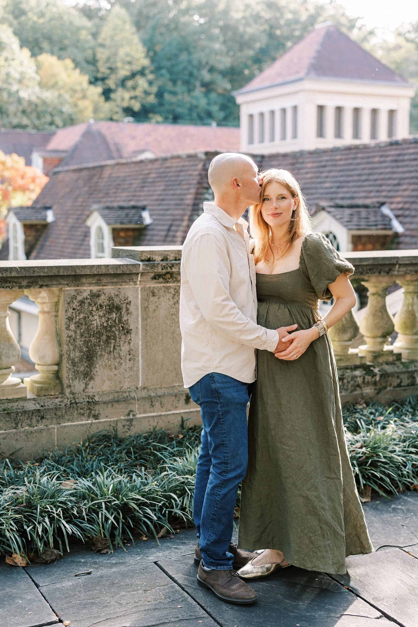 a pregnant couple stand together in golden hour light with beautiful Dupont architecture behind them at Winterthur