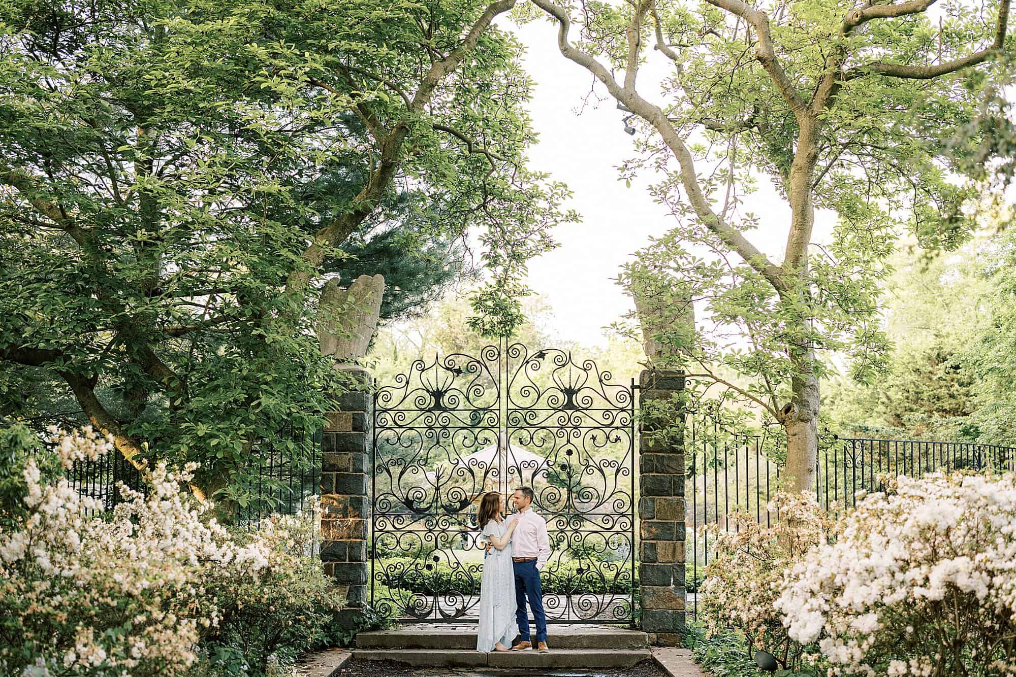 A couple stands holding each other in front of an ornate wrought iron gate. they are in a beautiful flowering garden at spring time called Marian Coffin Gardens at Gibraltar - a Dupont property