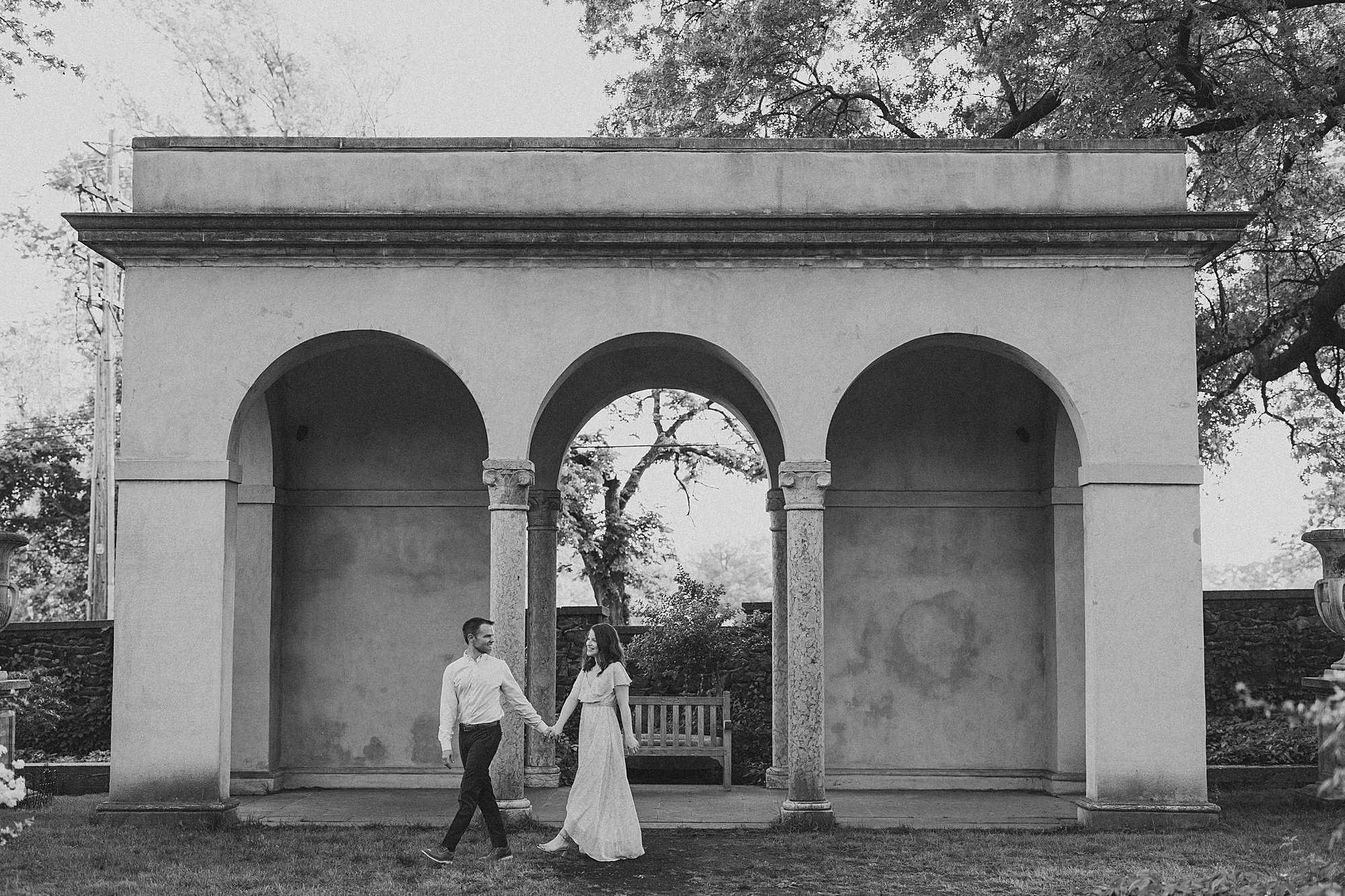 a black and white image of a groom leading his bride across a beautiful set of arches in a local park