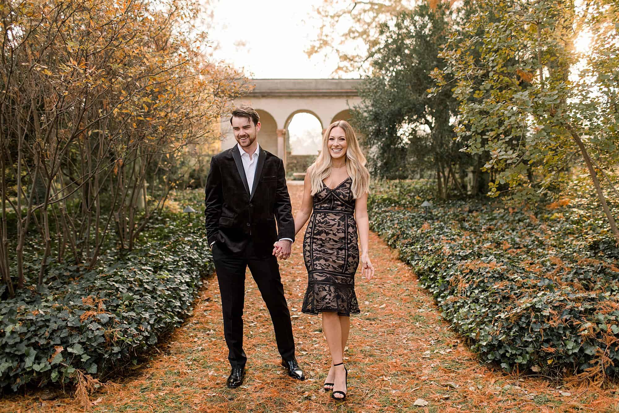 an engaged couple laugh together while holding hands in Marian Coffin Gardens at Gibraltar. he is wearing a black velvet suit and she is wearing a cocktail length black lace dress