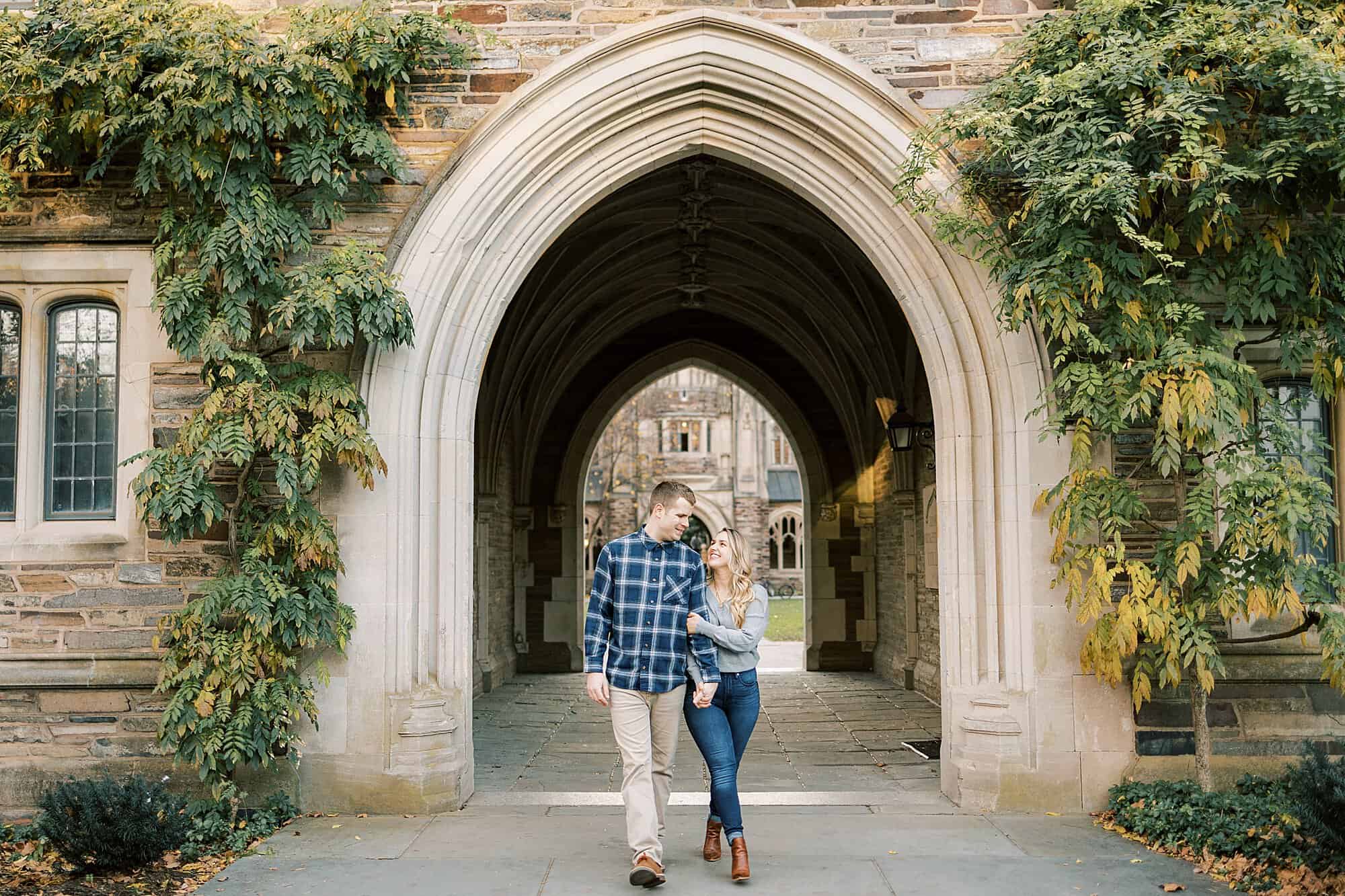 An engaged couple in casual clothes walk through the arches at Princeton University smiling and holding on to each other