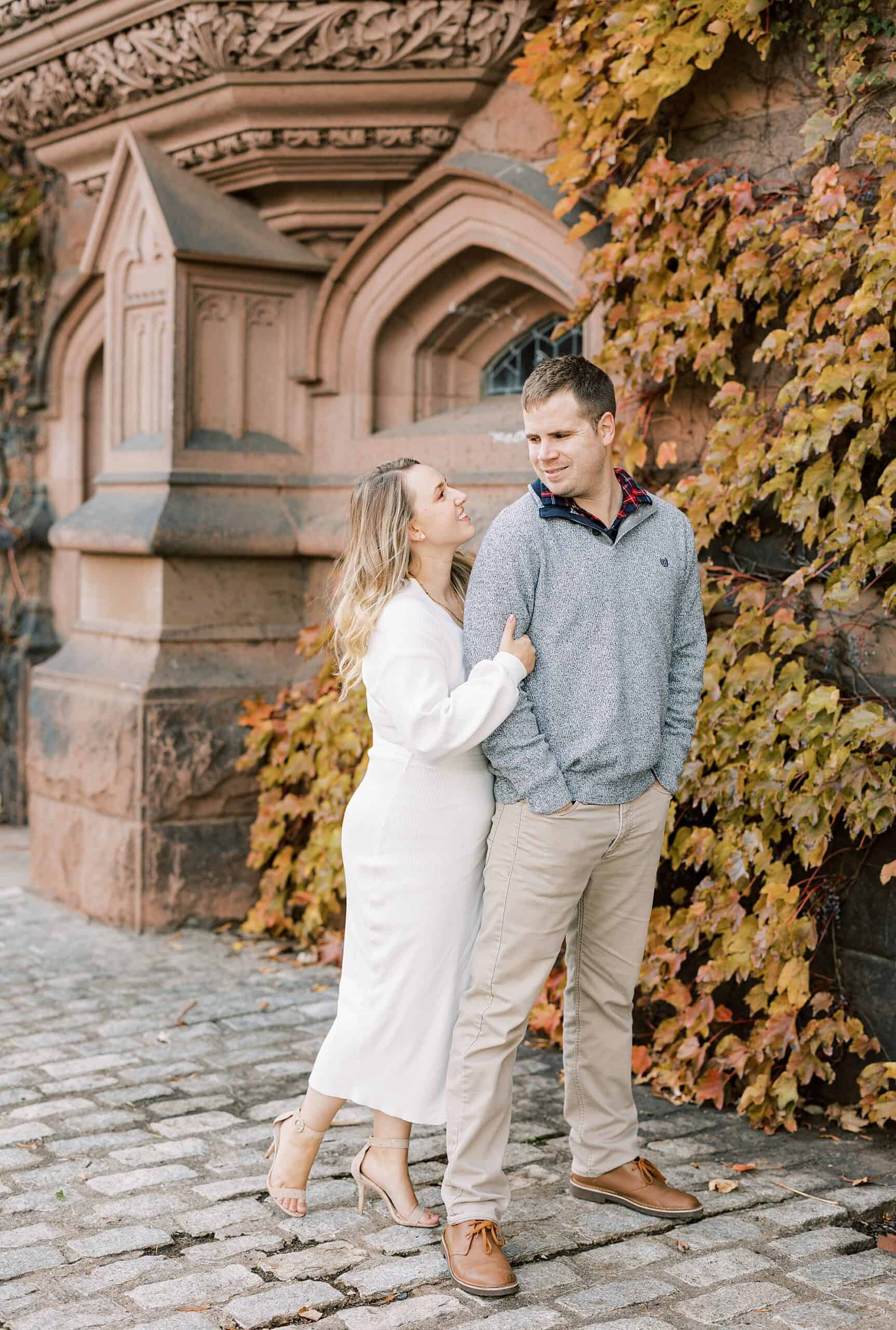 a woman in a white dress holds on to her fiance and smiles at him with surrounded by the beautiful architecture of Princeton University during fall