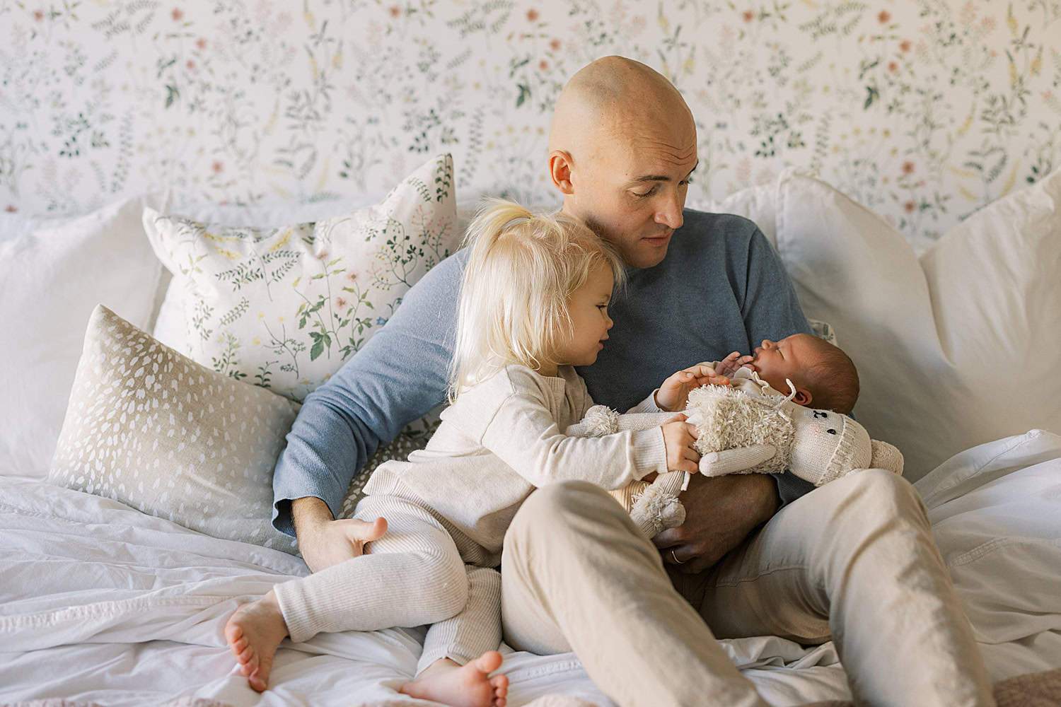 a father holds his newborn daughter and welcomes his toddler daughter on to his lap to gaze at the new baby during their in home newborn photos with Philadelphia newborn photographer Samantha Jay