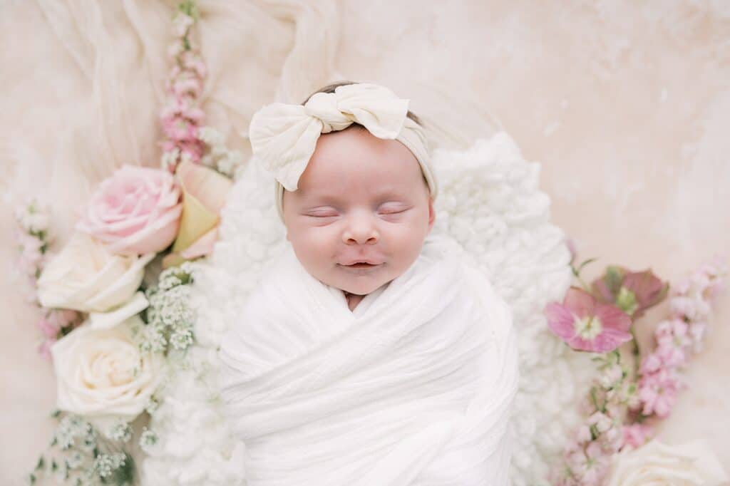 a fine art portrait of a newborn baby girl on a on a blush canvas backdrop surrounded by a floral halo of pink and cream fresh flowers during her newborn photoshoot with artist Samantha Jay 