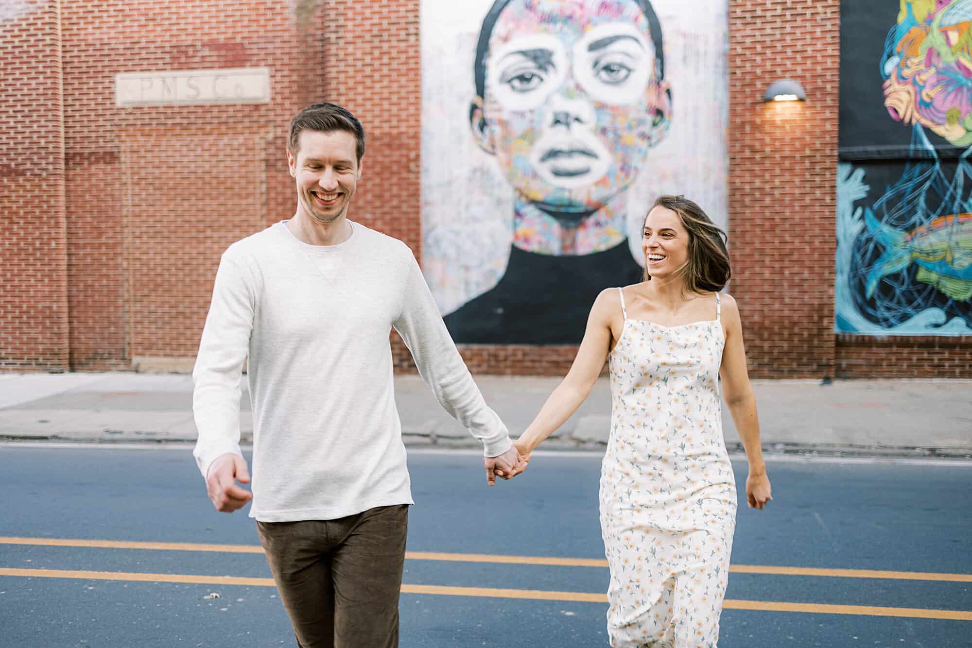 a handsome man and his beautiful fiance run across the streets of Philadelphia together in this candid photograph with a colorful Philadelphia mural behind them