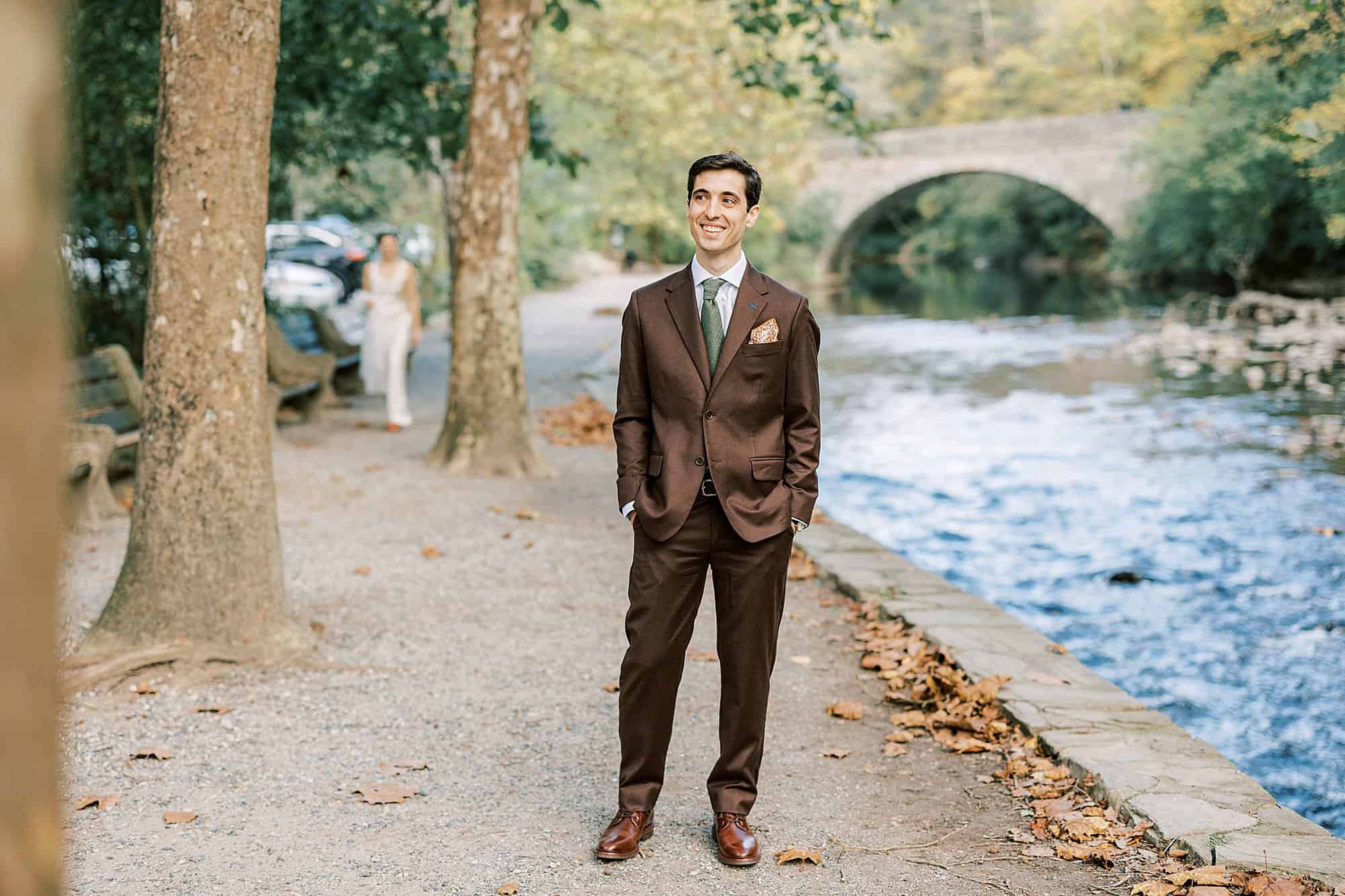 a groom in a brown suit waits for his bride next to a Wissahickon Creek at his Valley Green Inn wedding