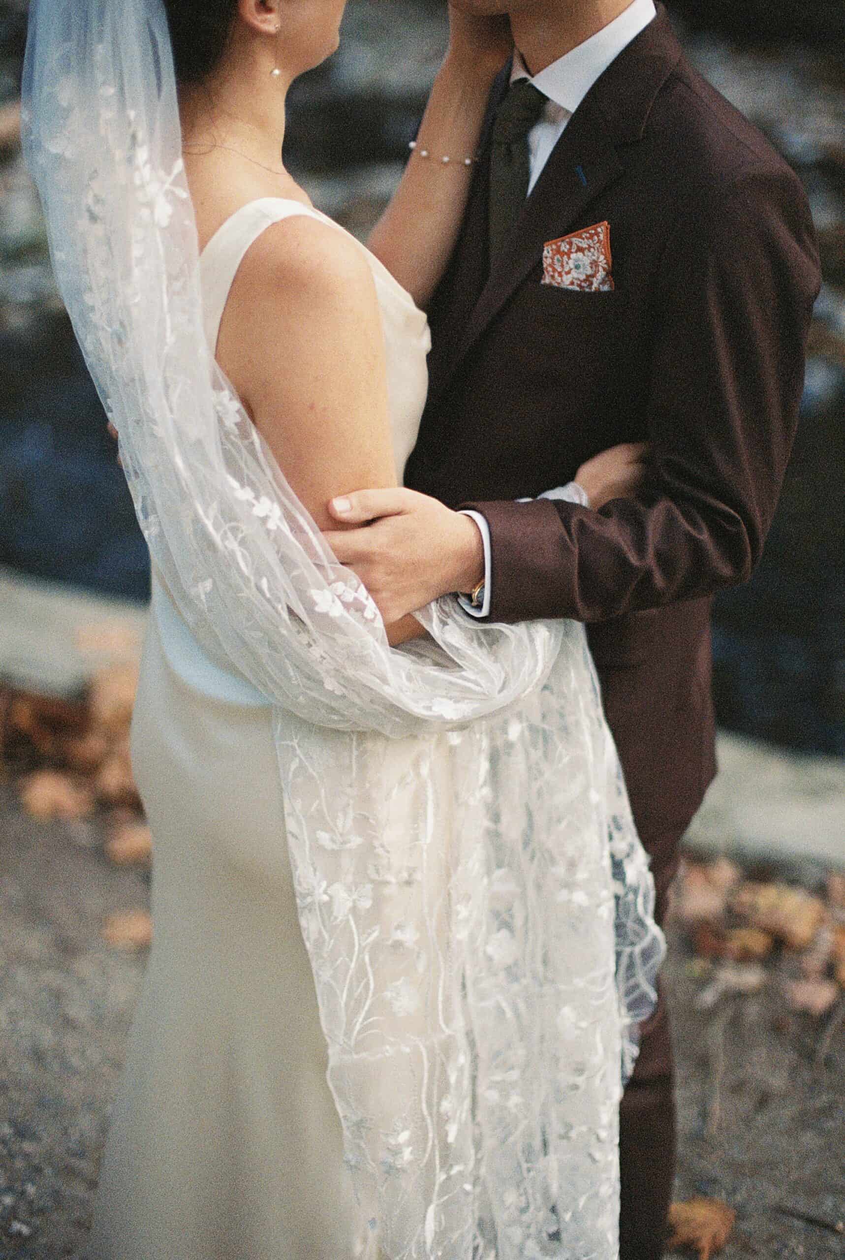 a film photography image of a Close-up of a bride and groom embracing outdoors, with the bride wearing a floral lace veil draped over her satin gown and the groom dressed in a dark suit with a patterned pocket square. The background features a natural setting with soft focus on a stream and fallen leaves