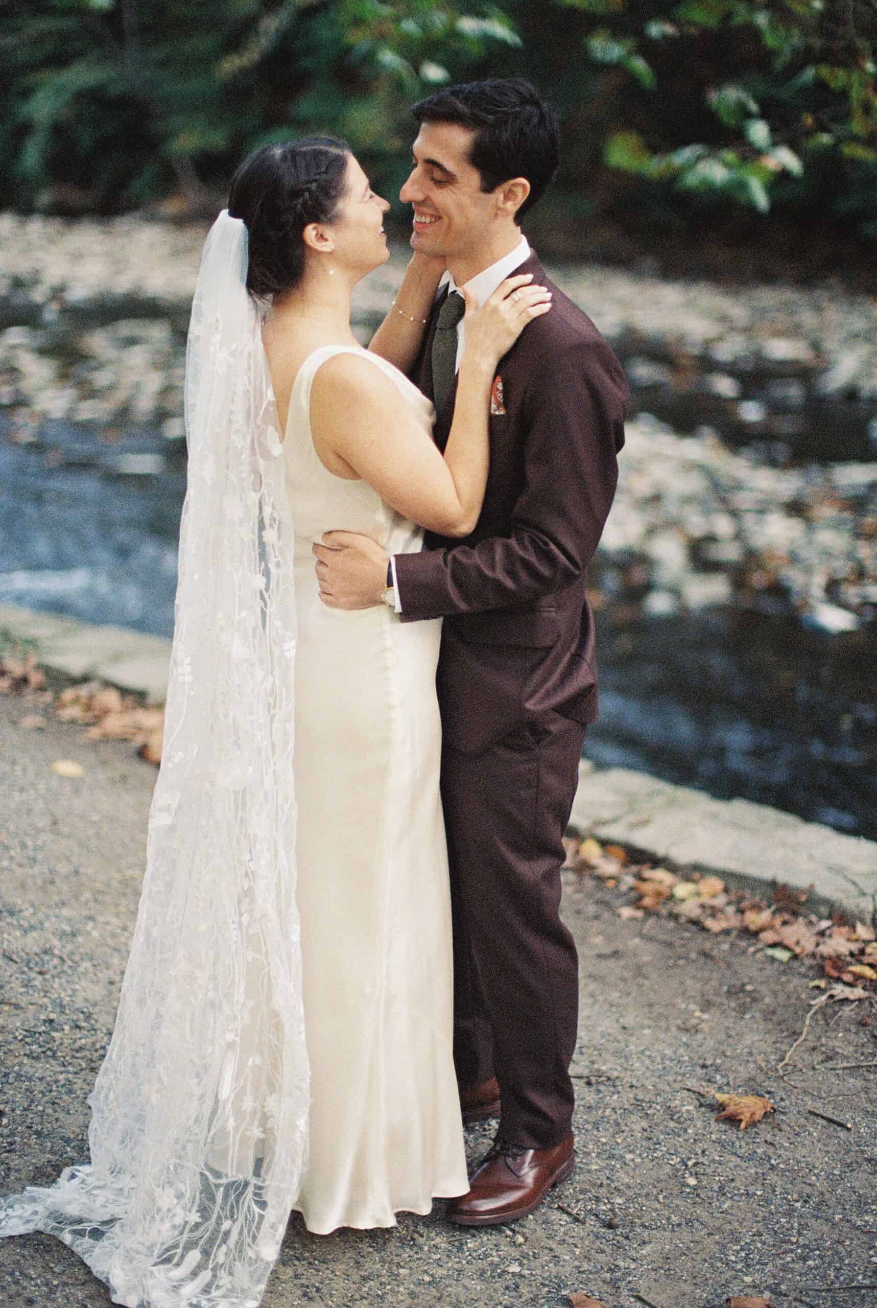 A bride and groom sharing a joyful moment outdoors by a serene stream. The bride wears a sleek satin gown and a floral lace veil, while the groom is dressed in a dark brown suit with a patterned pocket square. They stand on a gravel path surrounded by soft greenery and fallen leaves during their Valley Green Inn wedding