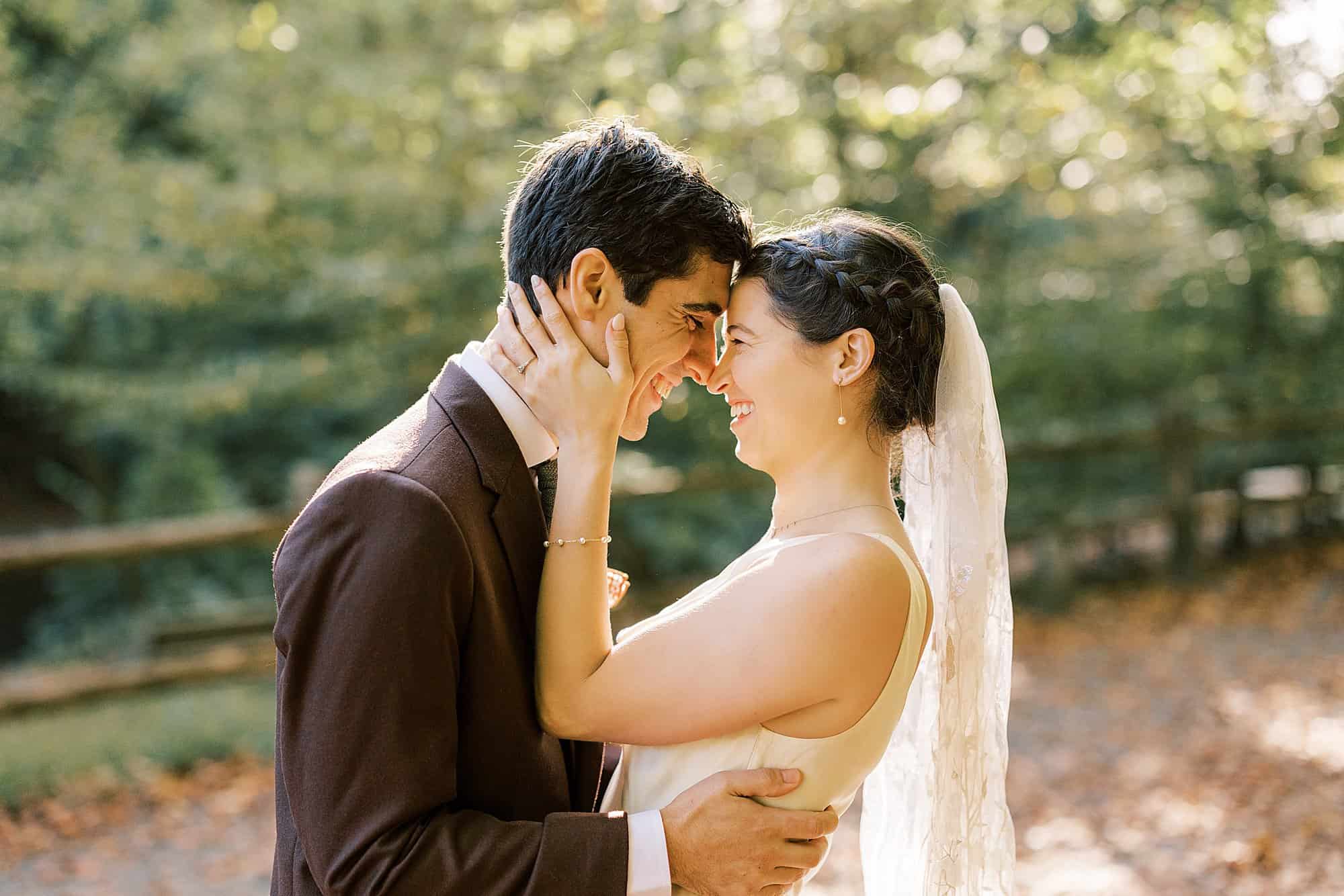 a bride wears a sleek satin gown and a floral lace veil holds her grooms face to hers as they laugh at each other