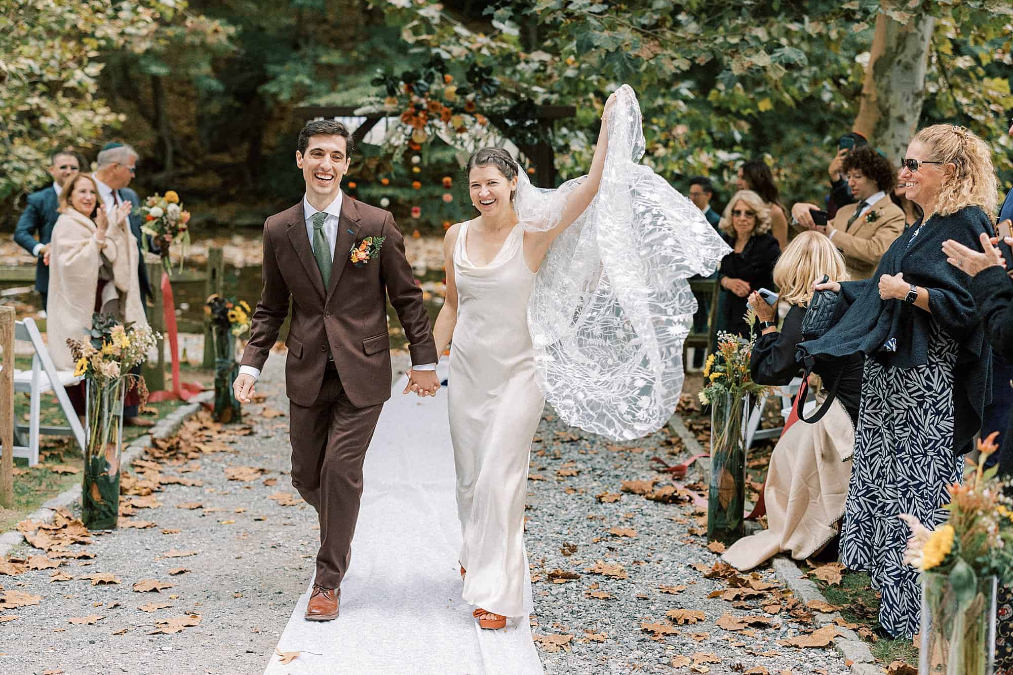 a bride waves her veil as she and husband recess down the aisle during their fall Valley Green Inn outdoor Jewish wedding ceremony 