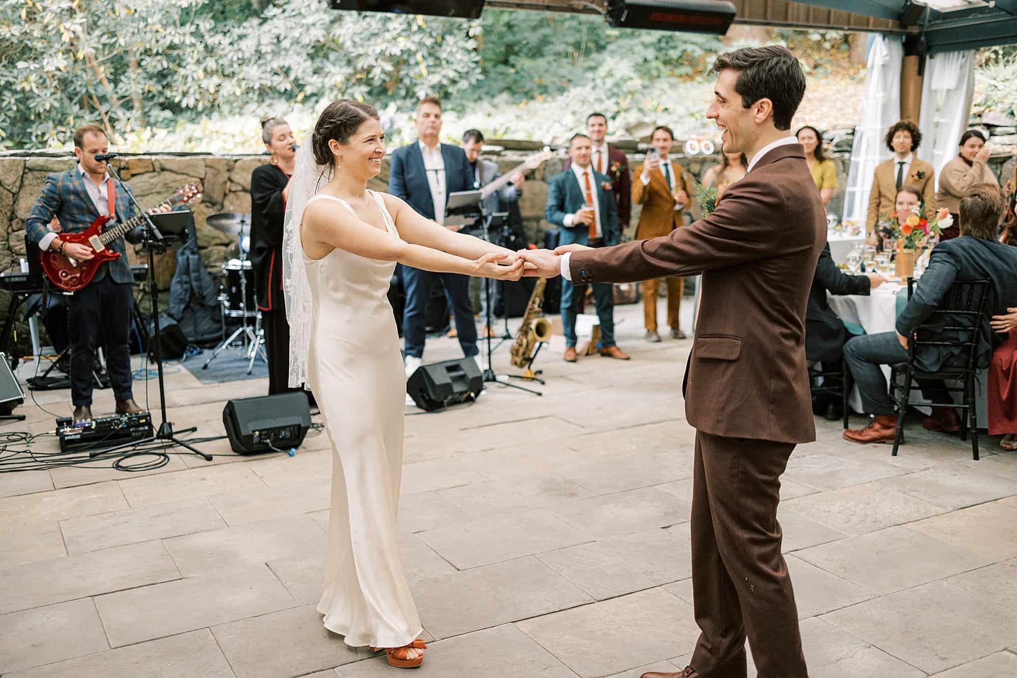 a bride and groom smile at each other while holding hands and dancing to their favorite song on their wedding day, a live band plays music behind them 