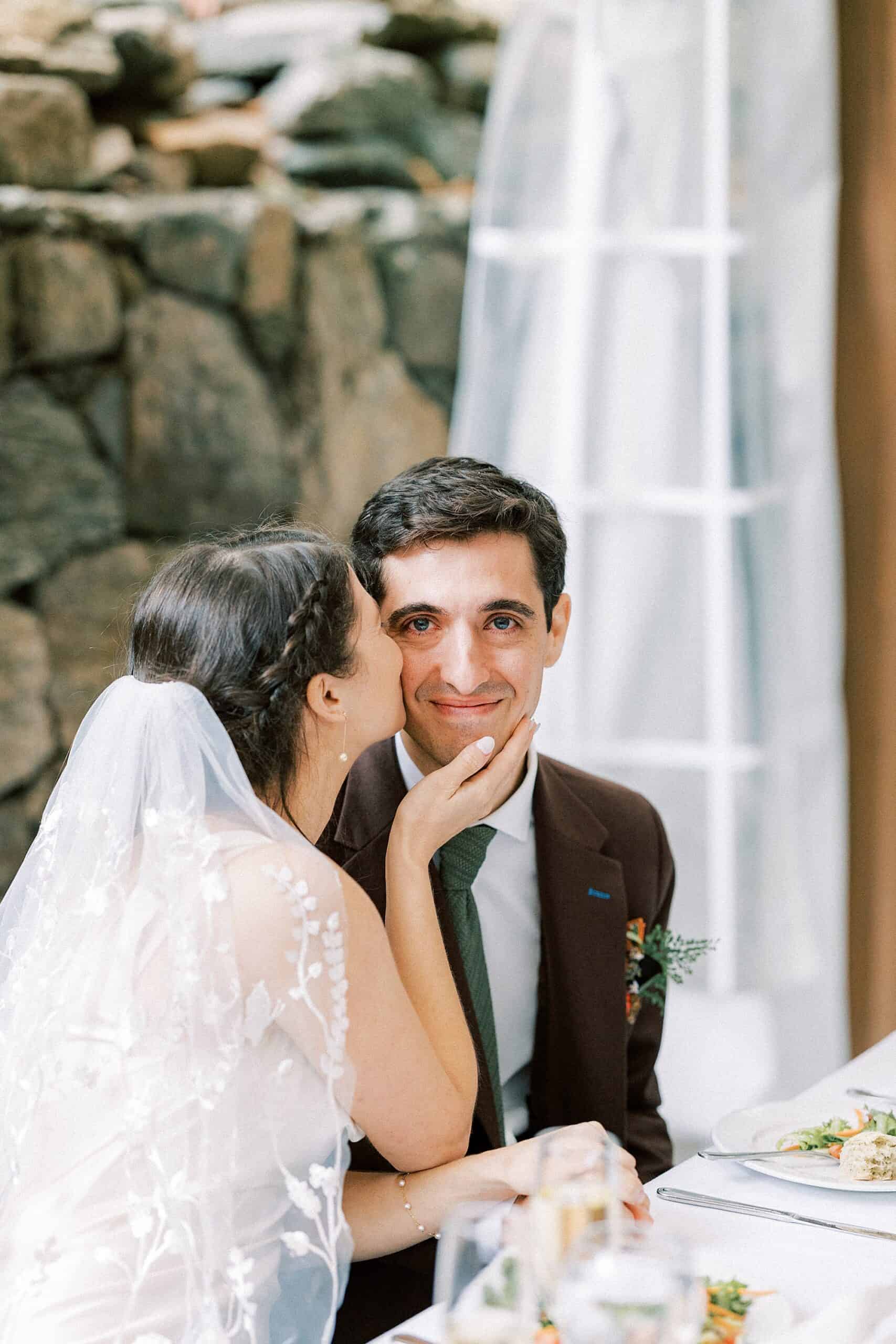 a groom in a custom brown suit smiles directly in the camera with tears in his eyes as his wife holds his cheek with her right hand and kisses his left cheek during their Valley Green Inn wedding reception