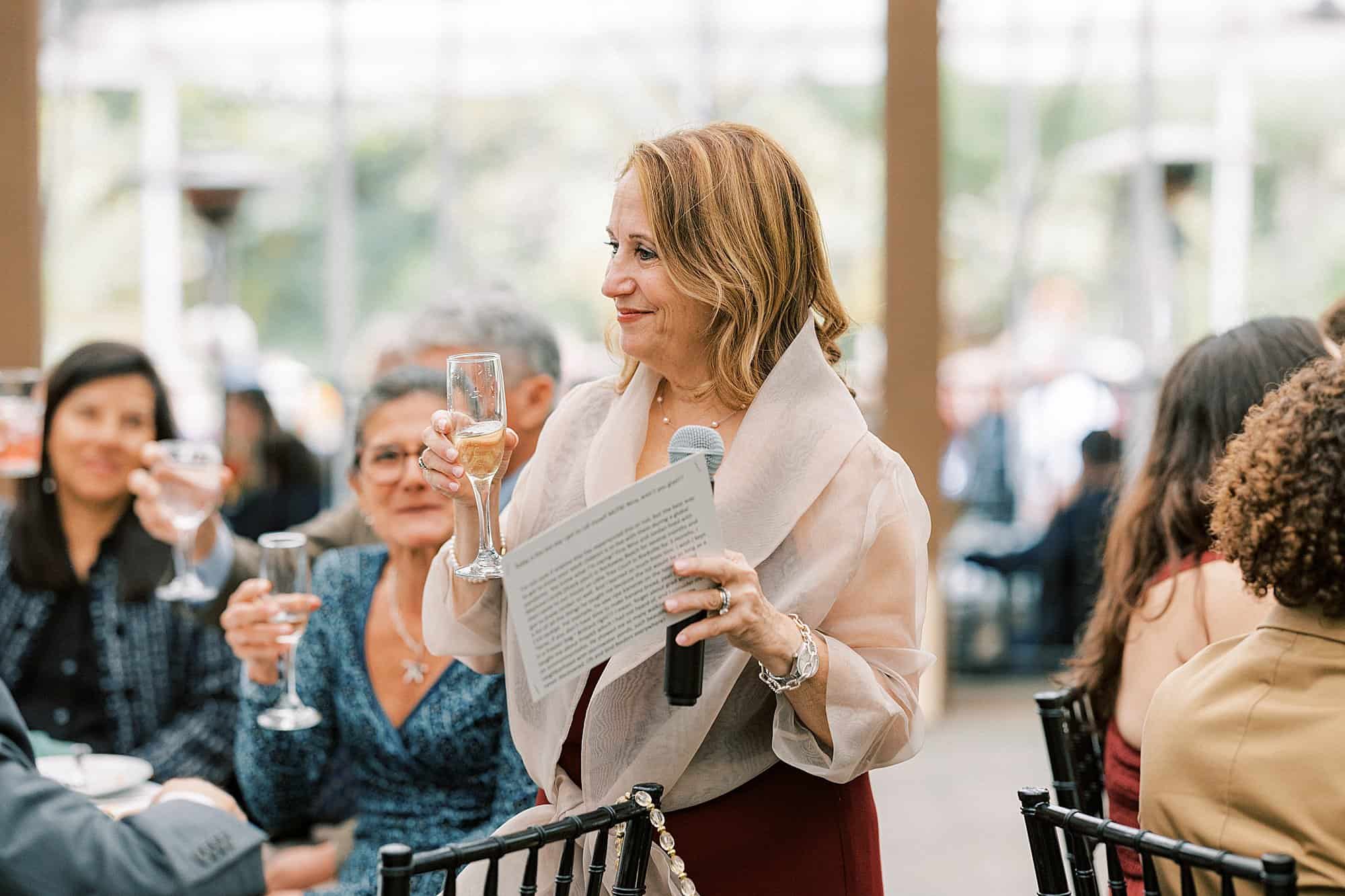 a mother holds a glass of champagne and a microphone and smiles while giving a toast at her daughters wedding