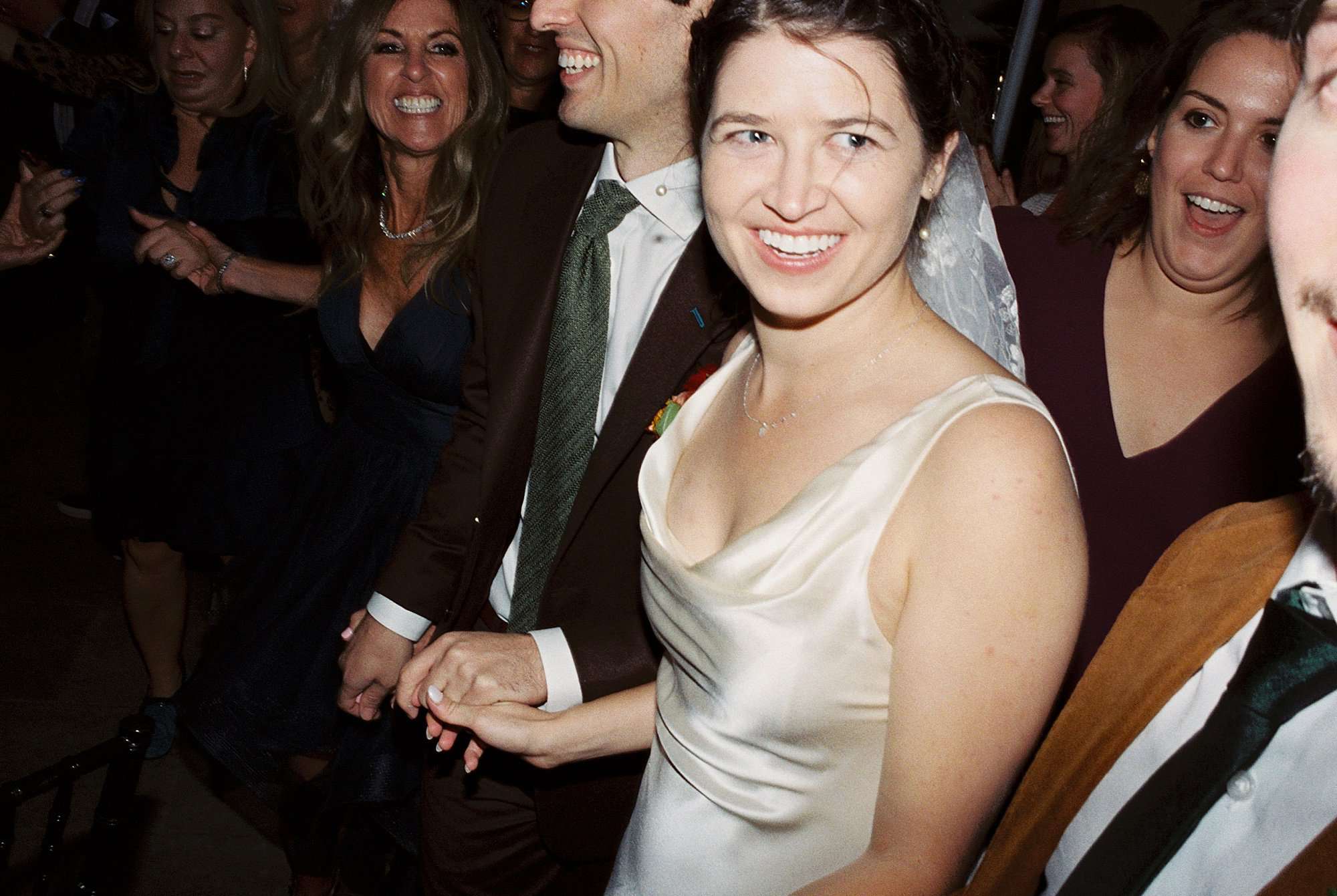 a candid film photography image of a bride smiling off camera surrounded by smiling guests while holding her husband's hand on the dance floor