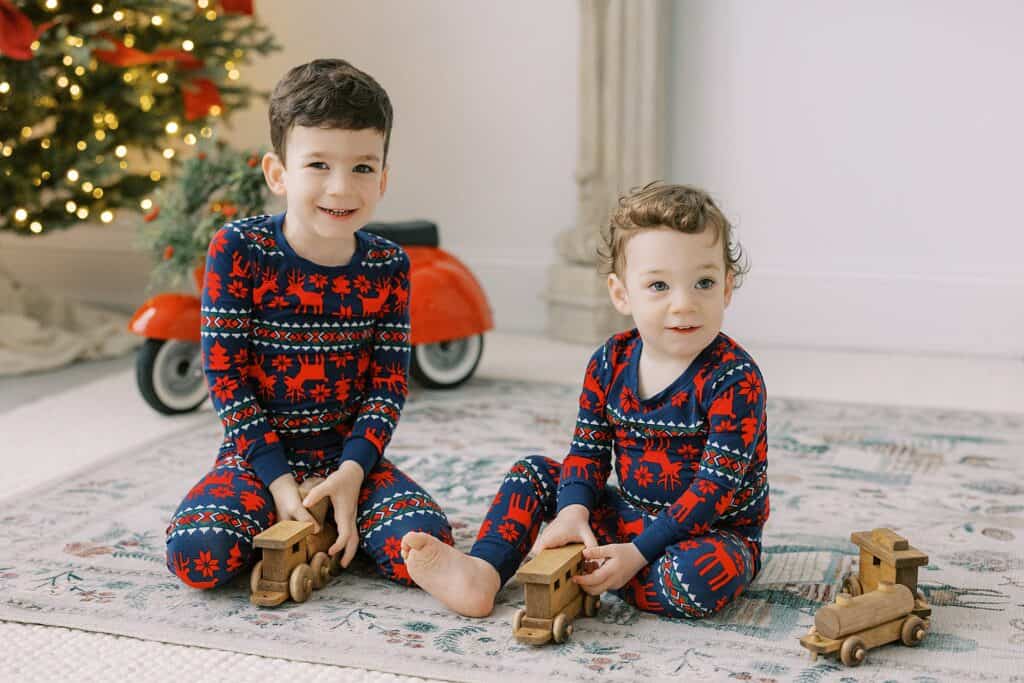 two brothers in Hanna Anderson pajamas play with a wooden train set surrounded by a Christmas tree and lots of joy