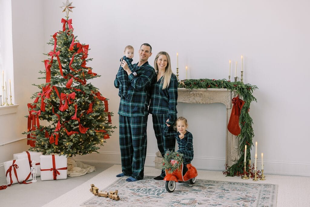a mother, father and their two daughters pose in their Christmas pajamas in front of a beautiful Christmas tree and mantle