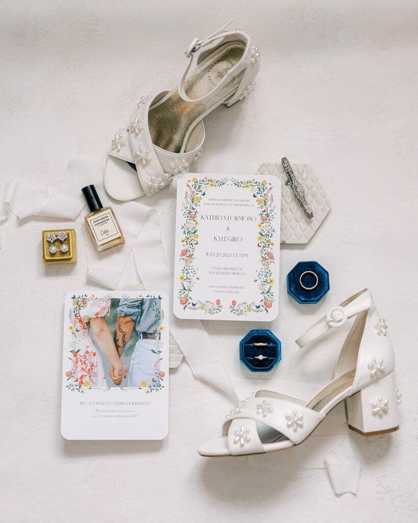 Flatlay of bridal details for a wedding at Tyler Arboretum in Philadelphia featuring white block-heel bridal shoes with pearl daisy appliqués, floral invitation suite, engagement rings in yellow and blue velvet ring boxes, pearl drop earrings, Curio perfume bottle, a vintage silver bracelet, silk ribbon, and a floral save-the-date card.