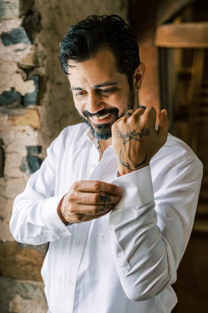 Groom smiling while buttoning his white dress shirt before his wedding at Tyler Arboretum in Philadelphia, with stone walls and warm natural light highlighting his tattooed hands.
