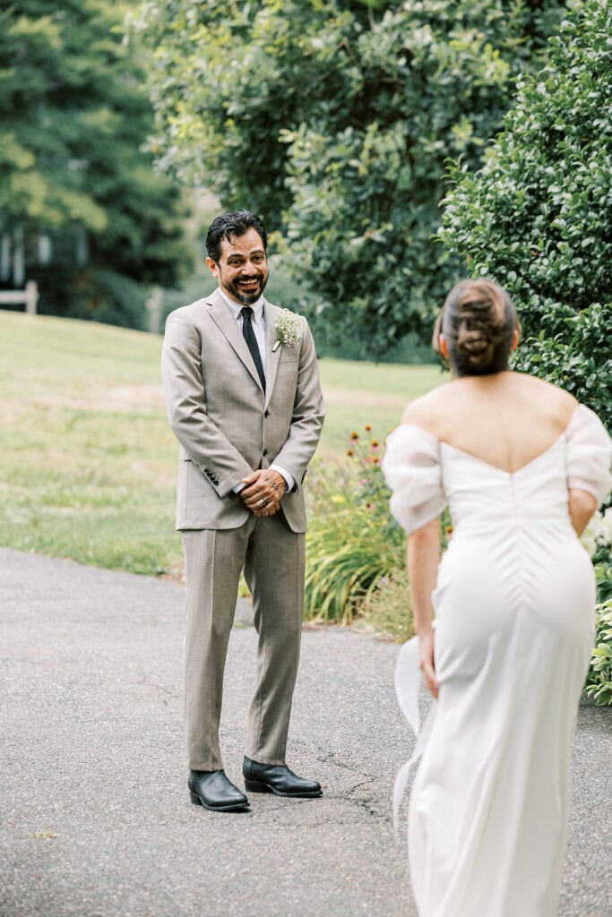 Groom smiling with anticipation as the bride approaches during their first look on a garden path at Tyler Arboretum in Philadelphia, wearing a tan suit with a baby’s breath boutonniere.