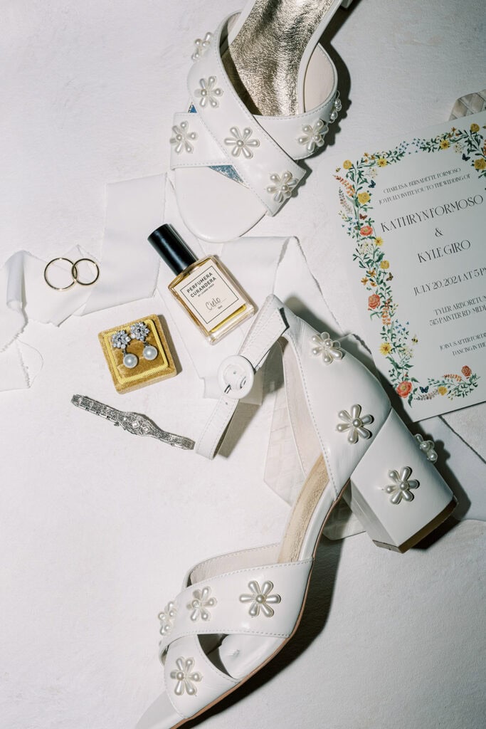 Close-up flatlay from a wedding at Tyler Arboretum in Philadelphia showcasing white daisy-embellished bridal heels, gold wedding bands, vintage pearl earrings, Curio perfume, floral wedding invitation, and a silver Art Deco bracelet styled against soft ivory fabric.