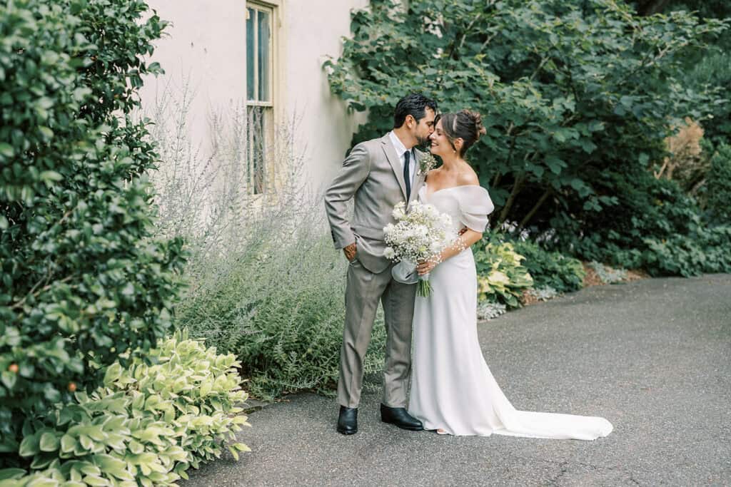 Bride and groom standing together beside a historic cream building and lush perennial gardens at Tyler Arboretum in Philadelphia, holding a bouquet of white daisies, chamomile, and baby’s breath during their wedding portraits.