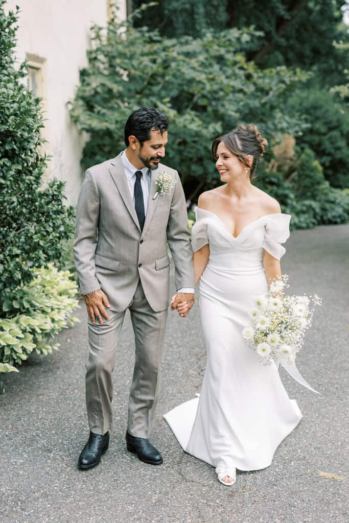 Bride and groom walking hand in hand down a shaded garden path at Tyler Arboretum in Philadelphia, showcasing the bride’s off-the-shoulder gown and her bouquet of white daisies, chamomile, and baby’s breath.