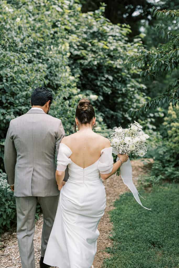 Bride and groom walking away from the camera along a wooded garden path at Tyler Arboretum in Philadelphia, the bride carrying a bouquet of white daisies, chamomile, and baby’s breath with long ribbon tails.