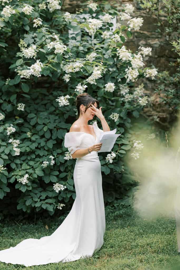 Bride reading her wedding vows beneath blooming white hydrangeas against a stone wall at Tyler Arboretum in Philadelphia, wiping away a tear during an emotional moment.