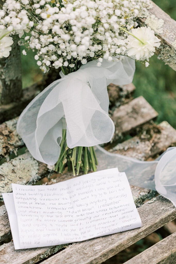 A folded sheet of white paper with handwritten wedding vows sits next to a bouquet of baby's breath tied with a sheer white ribbon on an aged wooden bench at Tyler Arboretum in Phialdelphia