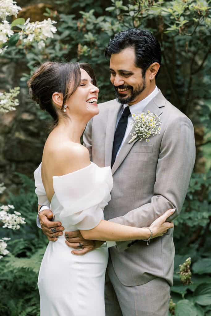 Bride and groom laughing and embracing in front of blooming hydrangeas during their wedding portraits at Tyler Arboretum in Philadelphia, with the groom wearing a tan suit and baby’s breath boutonniere.