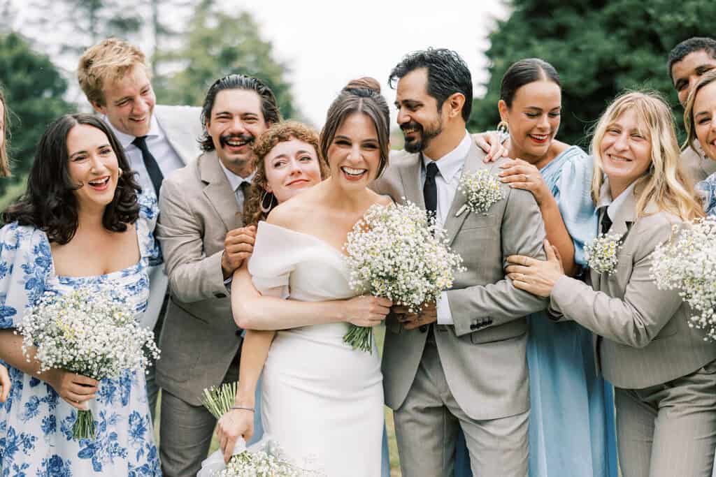 Bride and groom smiling with their wedding party on the lawn at Tyler Arboretum in Philadelphia, holding airy bouquets of baby’s breath and chamomile flowers, surrounded by bridesmaids in blue floral dresses and groomsmen in tan suits.