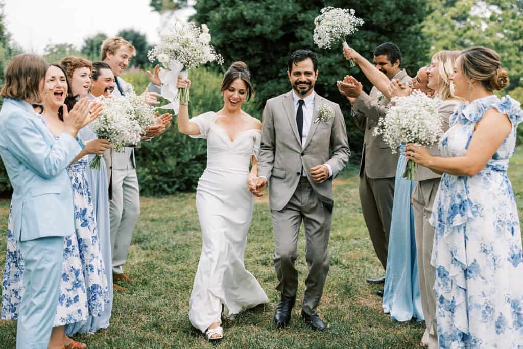 Bride and groom walking hand-in-hand through cheering wedding party at Tyler Arboretum in Philadelphia, holding white daisy, chamomile, and baby’s breath bouquets as bridesmaids in blue dresses celebrate around them.