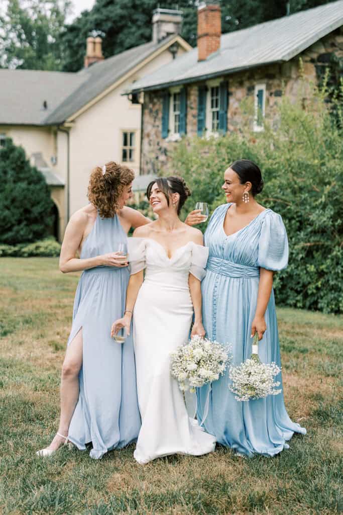 Bride laughing with her bridesmaids on the lawn at Tyler Arboretum in Philadelphia, holding glasses of wine and bouquets of baby’s breath and chamomile, with the venue’s historic stone house and lush gardens in the background.