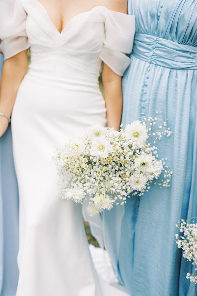 Close-up of the bride’s organic white bouquet featuring baby’s breath, white anemones, chamomile, and delicate white daisies held against her off-the-shoulder wedding gown at Tyler Arboretum in Philadelphia, beside bridesmaids in light blue dresses.